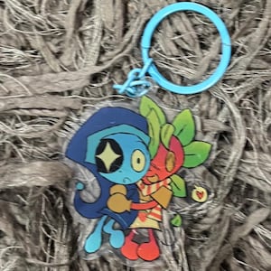 May include: A clear acrylic keychain featuring two cartoon characters embracing. One character is blue with a star-shaped eye, the other is red with green leaves. The keychain has a light blue ring.