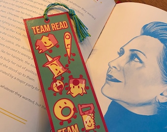 Op de afbeelding: Een groene bladwijzer met een rode rand heeft een cartoonfiguur met een glimlachend gezicht en de tekst "TEAM READ". De bladwijzer heeft ook een rode en gele cartoonzaag met een glimlachend gezicht en de tekst "TEAM 8".