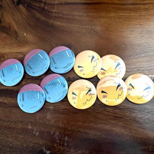 Puede incluir: Una colecci&oacute;n de diez chapas redondas con dos dise&ntilde;os distintos. Cinco chapas son de color azul claro con una parte superior rosa, y cinco chapas son amarillas con una ilustraci&oacute;n gr&aacute;fica de una cara sonriente con los ojos cerrados.
