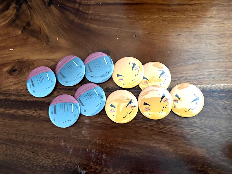 Puede incluir: Una colecci&oacute;n de diez chapas redondas con dos dise&ntilde;os distintos. Cinco chapas son de color azul claro con una parte superior rosa, y cinco chapas son amarillas con una ilustraci&oacute;n gr&aacute;fica de una cara sonriente con los ojos cerrados.