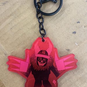May include: A red and pink keyring with a monster design. The keyring has a black chain and clasp. The monster has a skull face, a furry neck, and is wearing a headband. The design is on a red background.
