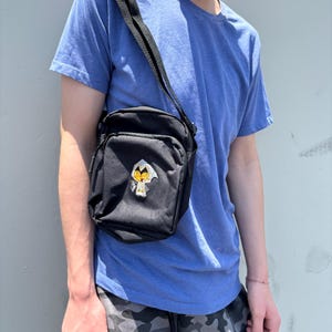 Pu&ograve; includere: Borsa a tracolla nera di forma rettangolare con una toppa ricamata. La borsa ha una tracolla nera regolabile. La persona nell'immagine indossa una maglietta blu e pantaloncini mimetici.