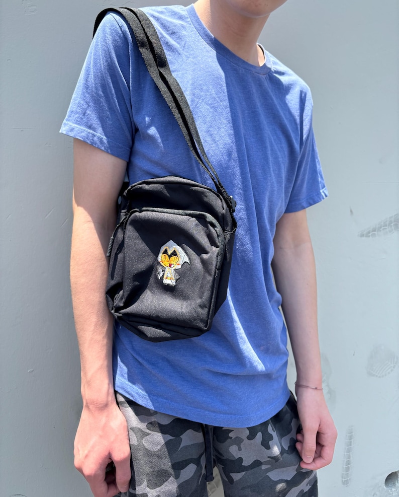Pu&ograve; includere: Borsa a tracolla nera di forma rettangolare con una toppa ricamata. La borsa ha una tracolla nera regolabile. La persona nell'immagine indossa una maglietta blu e pantaloncini mimetici.