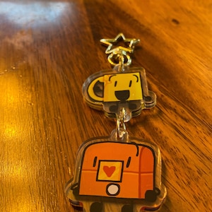 Puede incluir: Un llavero con dos dijes. El dije superior es una taza amarilla con una cara sonriente. El dije inferior es una caja naranja con un corazón en la parte delantera y un círculo blanco en el medio.