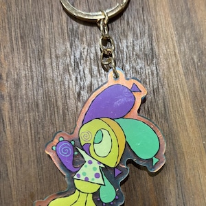 Puede incluir: Un llavero colorido con un personaje de dibujos animados con cuerpo amarillo, brazos verdes y cabeza morada. El personaje tiene una camisa de lunares y está unido a un llavero y cadena dorados. El fondo es una superficie de madera.