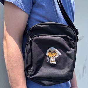 Pu&ograve; includere: Borsa a tracolla nera con toppa di personaggio dei cartoni animati. La borsa ha una forma rettangolare con una tasca frontale e una lunga tracolla regolabile. La toppa presenta un personaggio con cappuccio bianco e volto giallo.