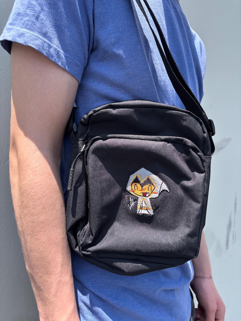 Pu&ograve; includere: Borsa a tracolla nera con toppa di personaggio dei cartoni animati. La borsa ha una forma rettangolare con una tasca frontale e una lunga tracolla regolabile. La toppa presenta un personaggio con cappuccio bianco e volto giallo.