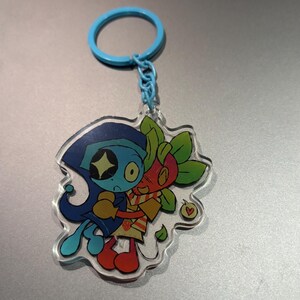 May include: A clear acrylic keychain featuring two cartoon characters embracing. One character is blue with a star-shaped eye, the other is red and green. The keychain has a blue ring and chain.