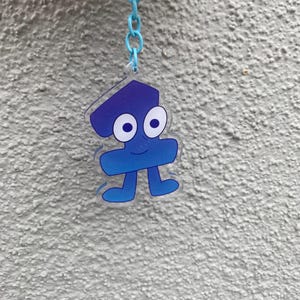 May include: A blue keychain featuring a cartoon character with a smiling face, large white-rimmed eyes, and small feet. The character is attached to a light blue chain and ring.
