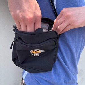 Pu&ograve; includere: Borsello nero con una piccola tasca frontale e chiusura a zip. La borsa presenta una grafica a cartoni animati sul davanti. Si indossa a tracolla con una tracolla nera.