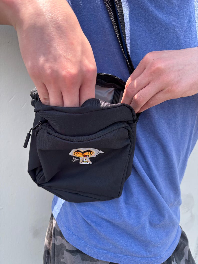 Pu&ograve; includere: Borsello nero con una piccola tasca frontale e chiusura a zip. La borsa presenta una grafica a cartoni animati sul davanti. Si indossa a tracolla con una tracolla nera.