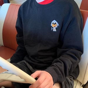 Puede incluir: Sudadera negra de cuello redondo con un pequeño dibujo animado en el pecho. El dibujo presenta un personaje con una capa con capucha. La sudadera tiene mangas largas y cuello y puños acanalados. Se lleva una camiseta roja debajo.