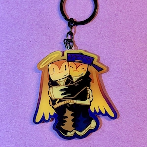 May include: A keychain featuring a cartoon illustration of two figures hugging. The figures have orange skin, black clothing, and are wearing a blue baseball cap. The keychain has a black ring and chain, and yellow and blue wings.
