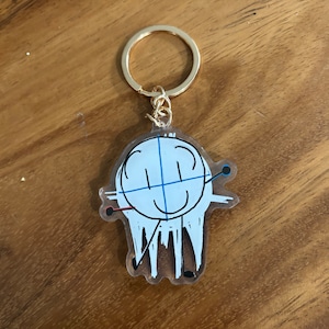 May include: A clear acrylic keychain featuring a whimsical cartoon figure with a white head, blue crosshairs, and a smiling face. The figure has black legs and arms with red and blue accents. The keychain has a gold-colored ring.
