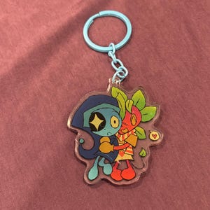 May include: A clear acrylic keychain featuring two cartoon characters hugging. One character is blue with a star-shaped eye, and the other is red with green leaves. The keychain has a light blue ring and chain.