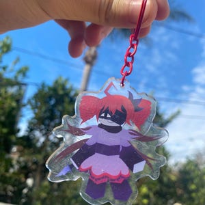 Puede incluir: Un llavero acrílico transparente con un personaje de dibujos animados con pelo rojo, una máscara negra y un atuendo morado y negro. El llavero tiene una cadena y un anillo rojos. El fondo muestra un cielo azul y follaje verde.