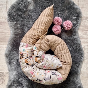 Könnte beinhalten: Ein beiges Baby-Nest mit Waffelstruktur und einem Innenmuster mit Tieren und einer hellbraunen Krawatte. Das Nest liegt auf einem grauen Kunstfellteppich, mit drei rosa Pomponblumen.