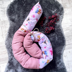 Könnte beinhalten: Ein Baby-Bettumrandung in Spiralform, die rosa Waffelstoff mit einem gemusterten Stoff mit Cartoon-Tieren, Regenbogen und Ballons kombiniert. Die Bettumrandung ist mit Bindfaden gebunden und liegt auf einem grauen, flauschigen Teppich.