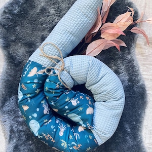 Könnte beinhalten: Ein Baby-Stützkissen mit blauem Waffelstoff und einem türkisfarbenen Stoff mit einem Meeresmuster. Das Kissen ist mit Bindfaden gebunden und liegt auf einer strukturierten, grauen Oberfläche. Rosa und braune Blätter sind im Hintergrund.