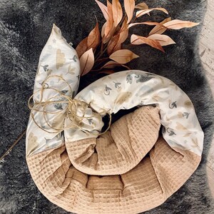 Könnte beinhalten: Ein beige-weißes Baby-Nest mit Waffelstruktur und einem bedruckten Design aus Wolken, Vögeln und Blättern. Das Nest ist aufgerollt und mit Schnur gebunden.