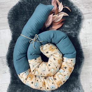 Könnte beinhalten: Ein blaues und cremefarbenes Baby-Nest mit Waffelstruktur und einem Muster aus Bären und Regenbögen. Das Nest ist zu einem Kreis zusammengerollt und liegt auf einem grauen, flauschigen Teppich.