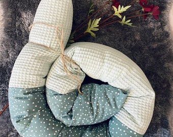May include: A long, cylindrical bolster pillow coiled into a spiral shape. One half features a light sage green waffle-textured fabric, while the other half is a darker sage green cotton with a white polka dot pattern, tied with thin twine.