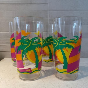 May include: Set of four clear glass drinking glasses with a tropical sunset design. The glasses feature a pink, orange, yellow, and green palm tree design with a sailboat.