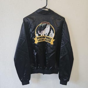 May include: Black satin bomber jacket with a full zip closure. The back features a circular embroidered patch with a wolf, trees, and the text "Nordik Wolf Light Beer". The jacket has a ribbed collar, cuffs, and hem.