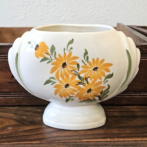 May include: An off-white ceramic vase with a wide, oval-shaped body and a pedestal base. The exterior features a hand-painted floral design with yellow daisy-like flowers, dark brown centers, and green leaves. The sides have vertical ribbed detailing.