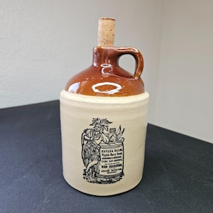 May include: A ceramic jug with a two-tone design featuring a glossy brown glazed top and a matte cream-colored base. A cork stopper is inserted into the neck. The front displays a black printed illustration of a person and text including Havana Balm.