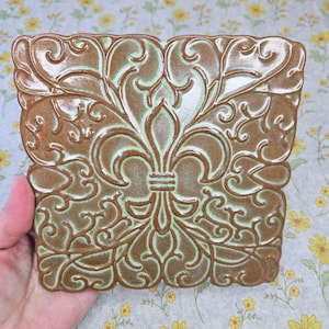 May include: A square ceramic tile featuring a raised, embossed fleur-de-lis design in the center. The surface has a glossy brown finish with light green accents highlighting the intricate, swirling scrollwork patterns across the entire piece.
