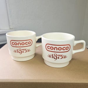 May include: Two off-white ceramic mugs with the Conoco logo in red, along with the year 1875. The mugs have a classic design with a handle and a stacked base. The mugs are sitting on a brown surface.