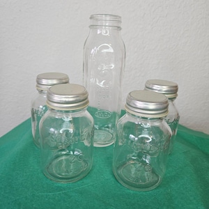May include: A collection of clear glass jars and bottles with silver metal lids. The jars have a rounded shape and are labeled with text. The bottles are of varying sizes, with one tall, rectangular bottle in the center.