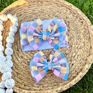May include: A set of two baby headbands with a patchwork design. The headbands feature a large bow with a mix of pastel colors, including pink, yellow, and blue, with a floral pattern. The headbands are placed on a woven, circular mat.