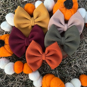 May include: A collection of fabric hair bows in various fall colors, including mustard yellow, burgundy, and rust orange. The bows are arranged with felt pumpkins and white and orange felt balls, creating a seasonal theme.