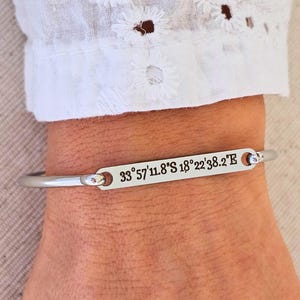 Può includere: Un bracciale a polsino color argento con coordinate incise: 33°57'11.8"S 18°22'38.2"E. Il bracciale è indossato su un polso, con una manica bianca ricamata con motivi traforati sullo sfondo. Il bracciale ha un design semplice ed elegante.
