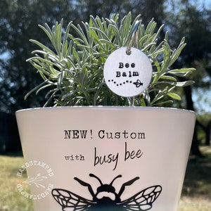 Op de afbeelding: Een witte keramische pot met een zwart bijenontwerp en de tekst "NEW! Custom with busy bee" aan de zijkant. Een zilveren metalen plantenmarker met de tekst "Bee Balm" en een klein bijenontwerp is bevestigd aan de plant.