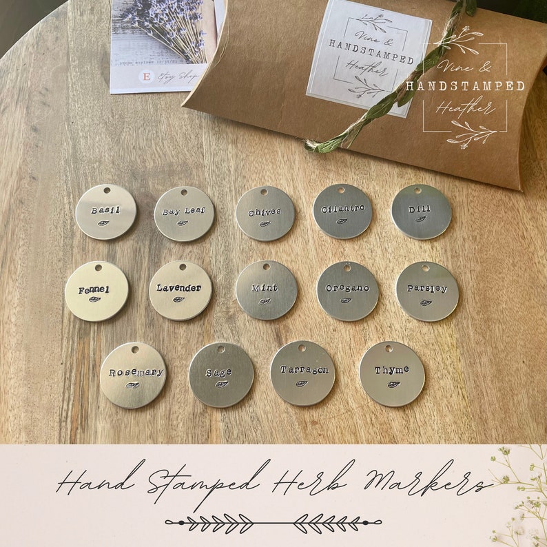 Op de afbeelding: Een set van 14 ronde zilveren metalen kruidenmarkeringen met handgestempelde namen van kruiden, waaronder basilicum, laurierblad, bieslook, koriander, dille, venkel, lavendel, munt, oregano, peterselie, rozemarijn, salie, dragon en tijm. De markeringen zijn in vier rijen op een houten oppervlak gerangschikt. De afbeelding bevat de tekst "Hand Stamped Herb Markers".