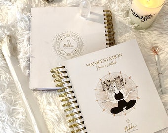 Può includere: Due quaderni bianchi a spirale con dettagli dorati. Uno presenta il testo "Manifestation Planner & Workbook" e un'illustrazione di una donna che medita. Sono visibili anche una candela accesa con "namaste" e mazzi di salvia.