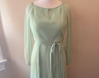 Könnte beinhalten: Ein langes, hellgrünes Plisseekleid mit langen Ärmeln und einem Bindegürtel. Das Kleid hat einen runden Ausschnitt und einen leicht gerafften Rock.