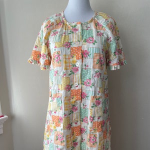 May include: A vintage patchwork robe with a floral design. The robe has a button-up front, short sleeves with frilled edges, and two pockets. The fabric is a mix of pastel colours, including pink, yellow, orange, and green, arranged in a square pattern.
