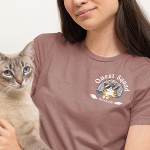 Pode incluir: Uma mulher vestindo uma camiseta malva com um gráfico de gato e o texto "Quest Squad Broccoli". Ela está segurando um gato cinza e branco em seus braços.