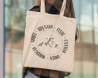 May include: A beige tote bag with a black circle design featuring a mountain range and stars. The circle is surrounded by the names "Azriel", "Morrigan", "Amren", "Cassian", "Feyre", "Rhysand".
