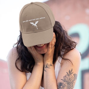 May include: A tan baseball cap with a white embroidered dragon and the text "we do not eat our allies".