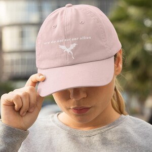 May include: A pink baseball cap with a white embroidered dragon and the text "we do not eat our allies".