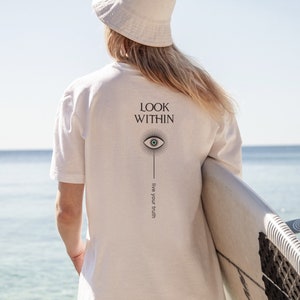 May include: White t-shirt with the text "LOOK WITHIN" and a stylized eye graphic with the text "live your truth" below. The person is wearing a white bucket hat and holding a surfboard.