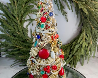 May include: A handcrafted Christmas tree sculpture, adorned with a variety of vintage jewelry pieces. The tree features red, green, blue, and pearl accents, with gold-toned metal elements. The tree is displayed on a mirrored surface, with a wreath in the background.