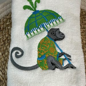 May include: Embroidered kitchen towel featuring a monkey wearing a green and gold patterned robe, holding a green and blue umbrella with a white fringe.