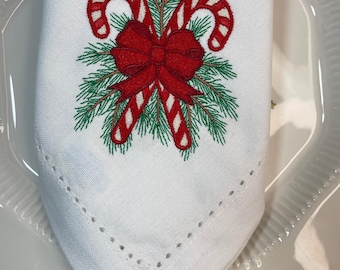 Op de afbeelding: Witte stoffen servet met borduursel van kerstkransjes en strik. Het ontwerp toont twee rood-witte zuurstokken, een rode strik en groene dennennaalden. Het servet ligt op een wit bord met een groene rand.
