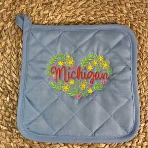 Op de afbeelding: Blauwe, gestikte ovenwant met een hartvormig ontwerp met een krans van bloemen en het woord "Michigan" geborduurd in rood.